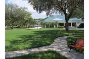 A well-maintained outdoor area at Sunnyside Village featuring a curved concrete walkway, green grass, mature trees providing shade, a red bench, and a building with a green roof in the background under a clear sky.