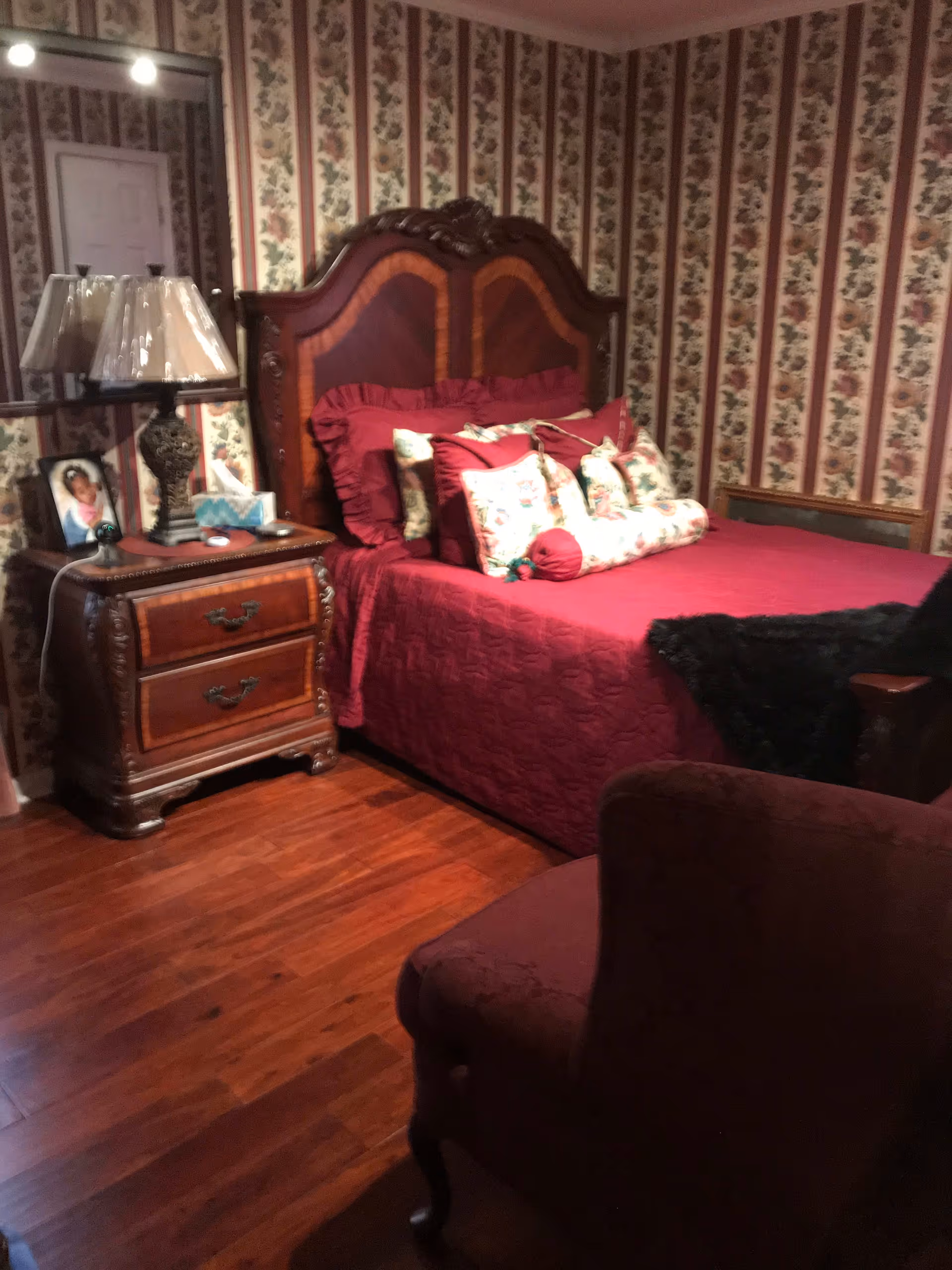 A bedroom with a wooden bed frame and headboard, covered with a red quilt and multiple decorative pillows. Next to the bed is a wooden nightstand with two drawers, a table lamp, a framed photo, and some tissues. The walls have floral wallpaper with vertical stripes. A mirror is mounted on the wall behind the nightstand, reflecting a white door. The floor is wooden, and a red upholstered chair is partially visible in the foreground.