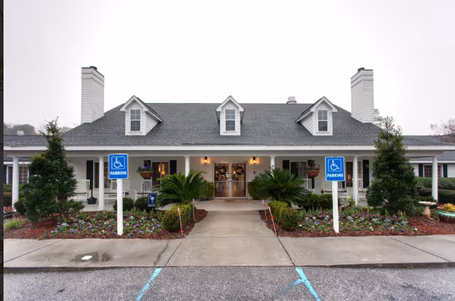Front exterior view of Cooper River Villages building with a gray roof, white walls, three dormer windows, two chimneys, and a covered porch. There are two handicap parking signs and landscaped flower beds with shrubs and small trees in front.