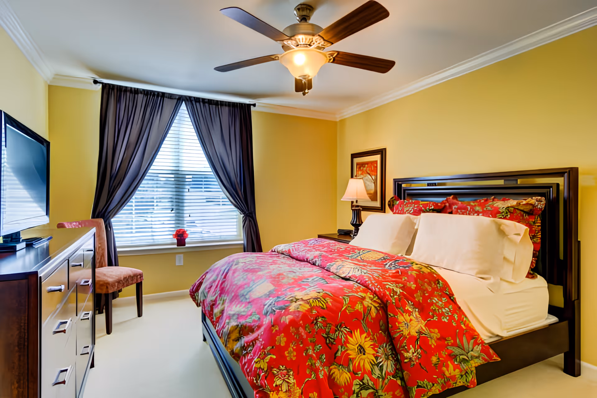 A cozy bedroom with yellow walls, a large bed with a colorful red floral comforter and white pillows, a wooden dresser with a flat-screen TV on top, a brown upholstered chair, a window with dark curtains, a bedside table with a lamp, and a framed picture on the wall.