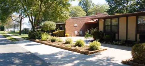 Brick assisted living building with front entrance, landscaped shrubs, and shaded walkway.