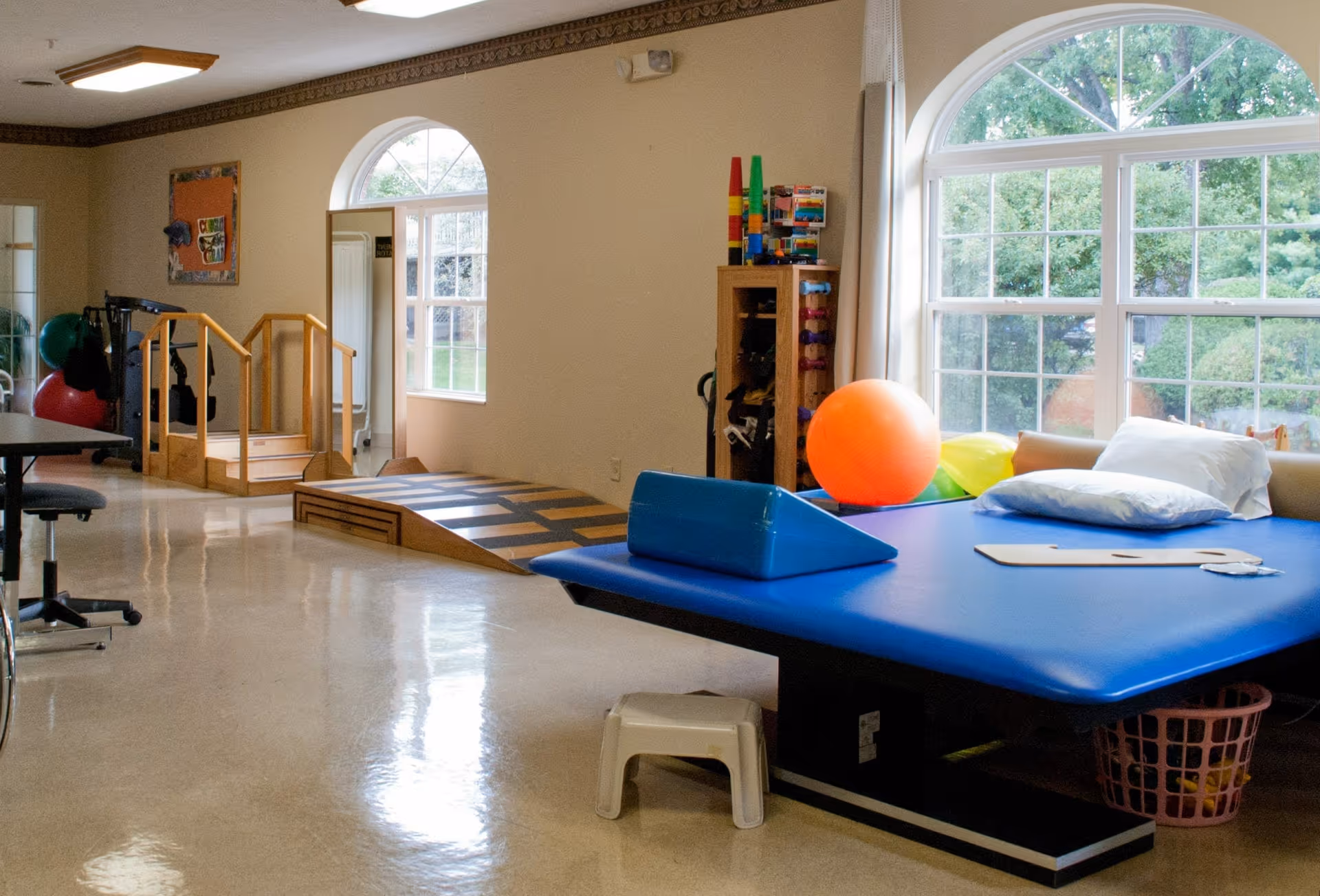 A bright therapy or rehabilitation room with large arched windows letting in natural light. The room contains a blue therapy table with pillows and exercise equipment including an orange exercise ball, small weights, and a wooden ramp with handrails. There is a mirror on the wall and a bulletin board with colorful papers.