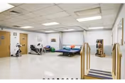Bright rehabilitation room with therapy tables, parallel bars, stationary bikes and exercise equipment.