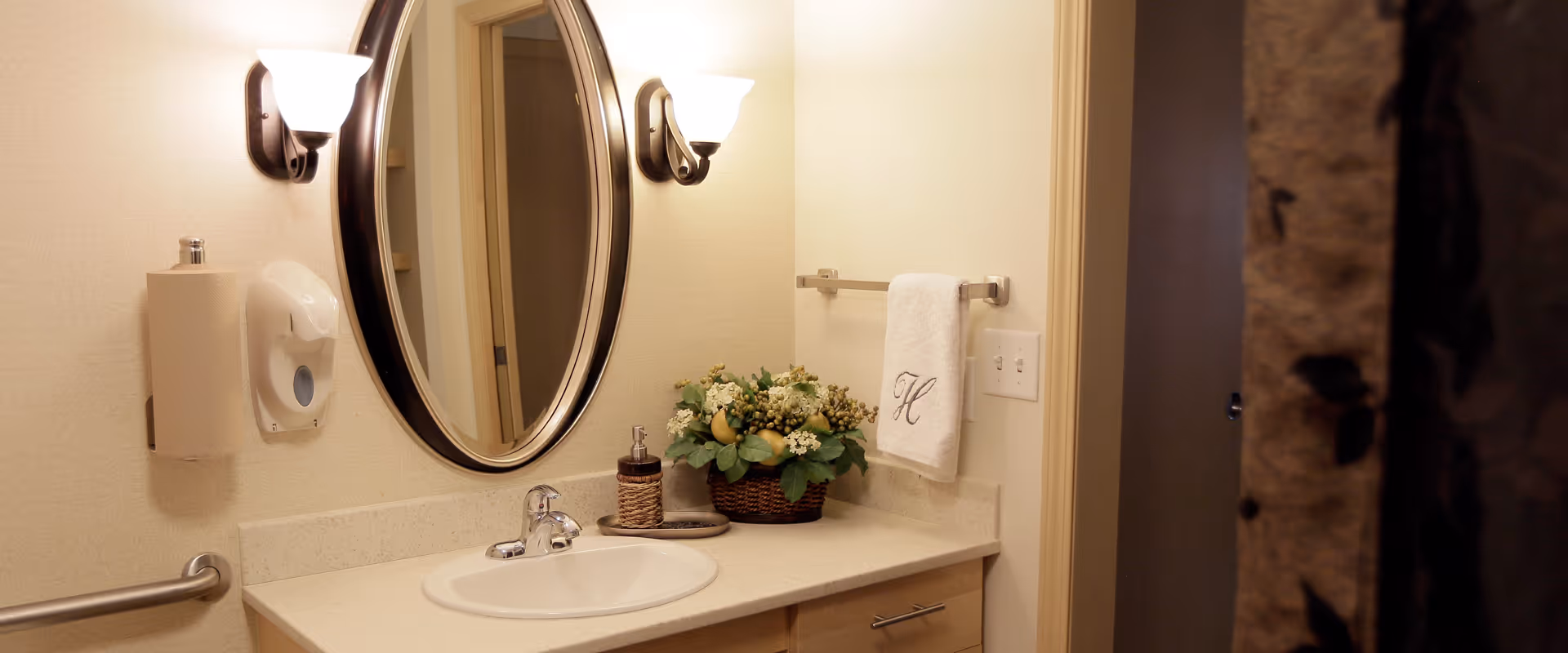 A bathroom vanity area with a white sink, a large oval mirror with a dark frame, two wall-mounted light fixtures, a soap dispenser, a paper towel holder, a hand towel with the letter 'H' embroidered on it, and a small basket of artificial flowers on the countertop.