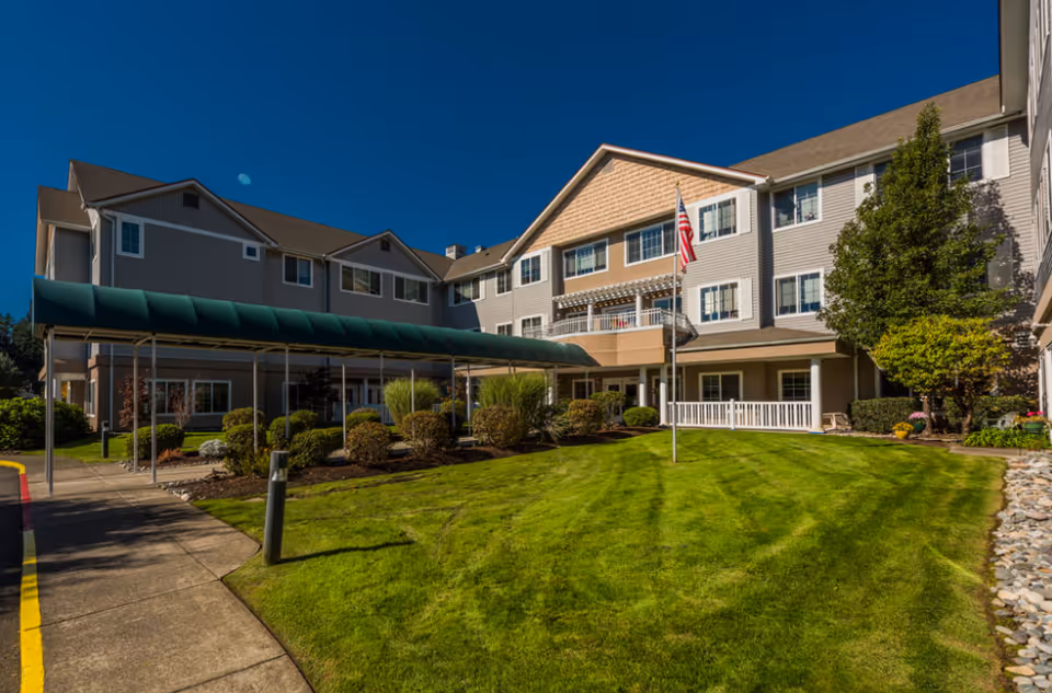 Front exterior of a multi-story senior living building with a covered entrance walkway, American flag, and manicured lawn.