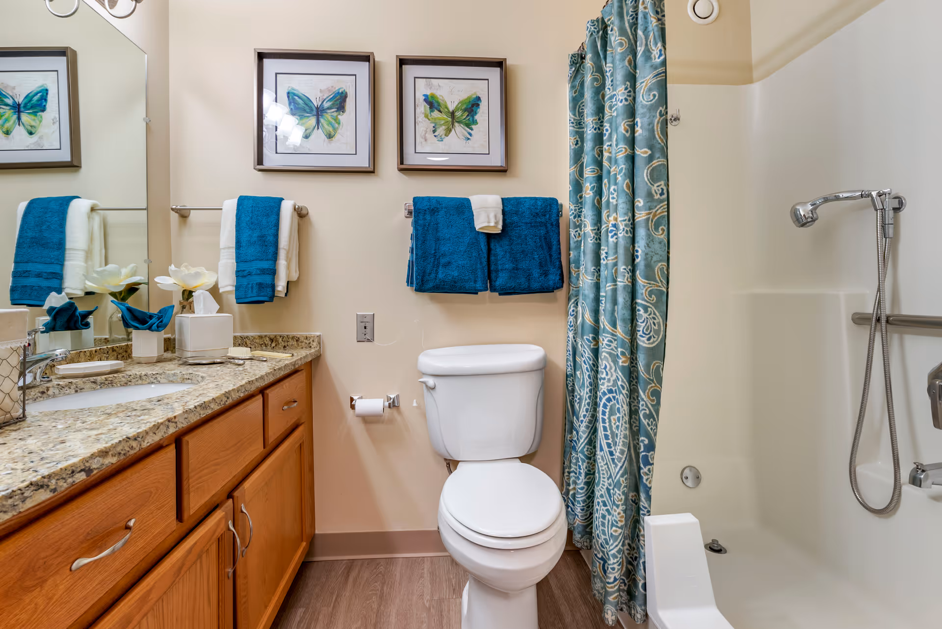 A clean and well-lit bathroom featuring a white toilet, a granite countertop with a sink, wooden cabinets, and a shower with a blue patterned curtain. Two framed butterfly artworks hang on the wall above blue and white towels.