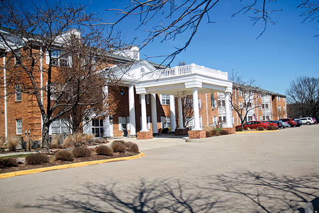 Brick senior living building with a white columned entrance canopy, parked cars, and leafless trees under a clear blue sky.