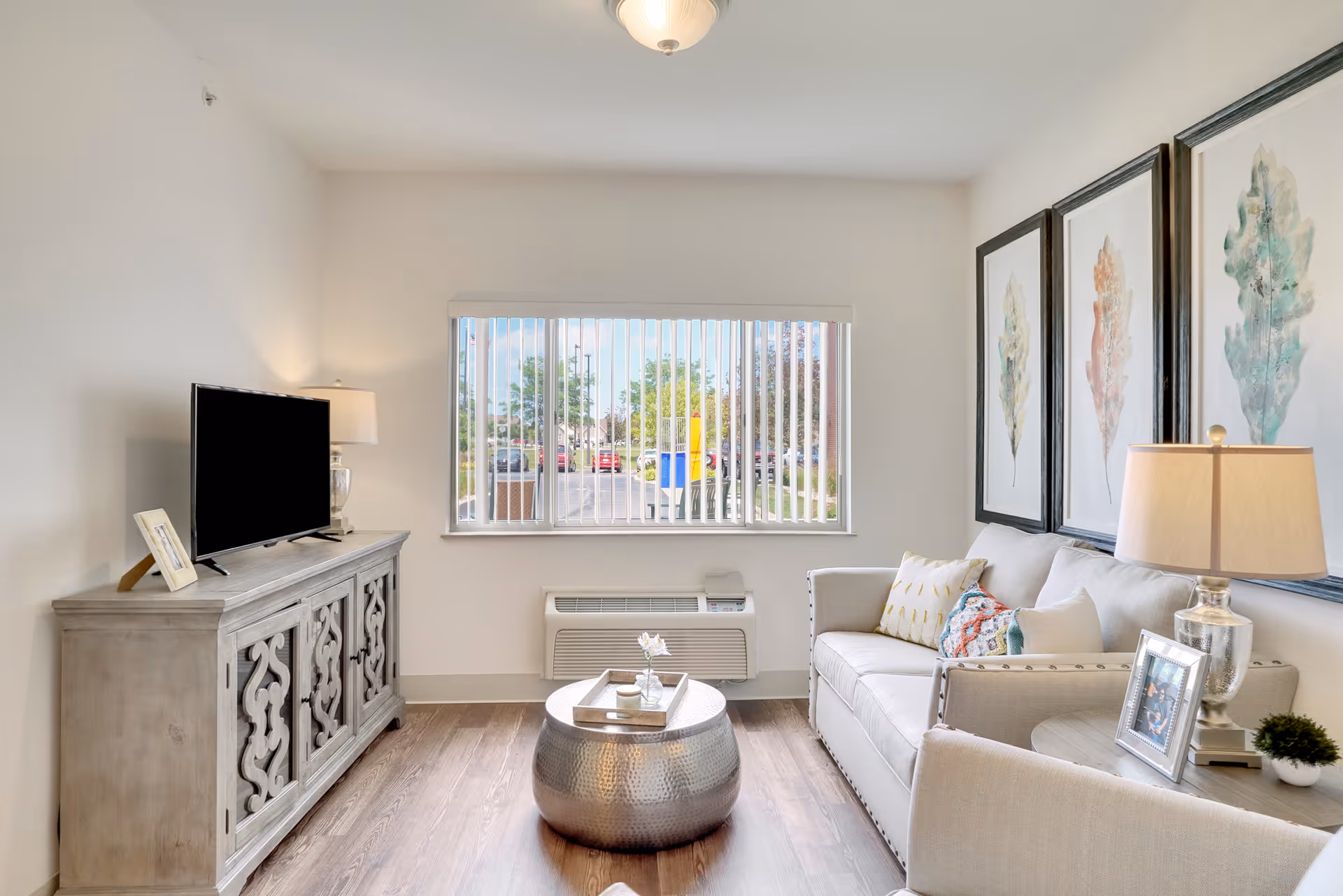 A bright living room with a large window featuring vertical blinds. The room includes a beige sofa with decorative pillows, a round metallic coffee table with a tray and flower vase, a side table with a lamp and framed photo, and a wooden TV stand with a flat-screen TV and a lamp. Three framed leaf artworks hang on the wall above the sofa.