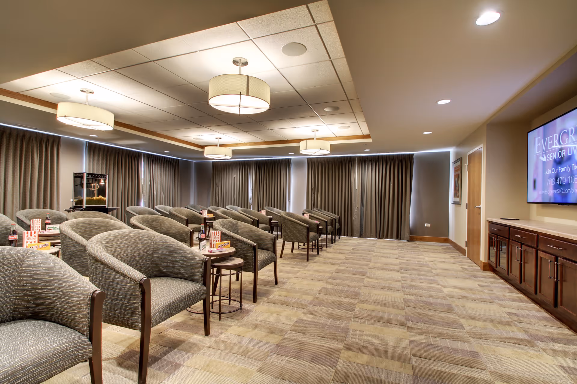 A spacious room with multiple rows of cushioned chairs arranged facing a large flat-screen TV mounted on the wall. The room has carpeted flooring, ceiling lights with round fixtures, and large windows covered by dark curtains. Small tables between chairs hold snacks and drinks, and a popcorn machine is visible in the back corner.