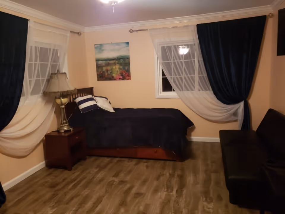 A bedroom with a single bed covered in dark bedding and two pillows, a wooden nightstand with a decorative lamp, two windows with sheer white and dark blue curtains, a painting on the wall, and a black sofa on the right side.