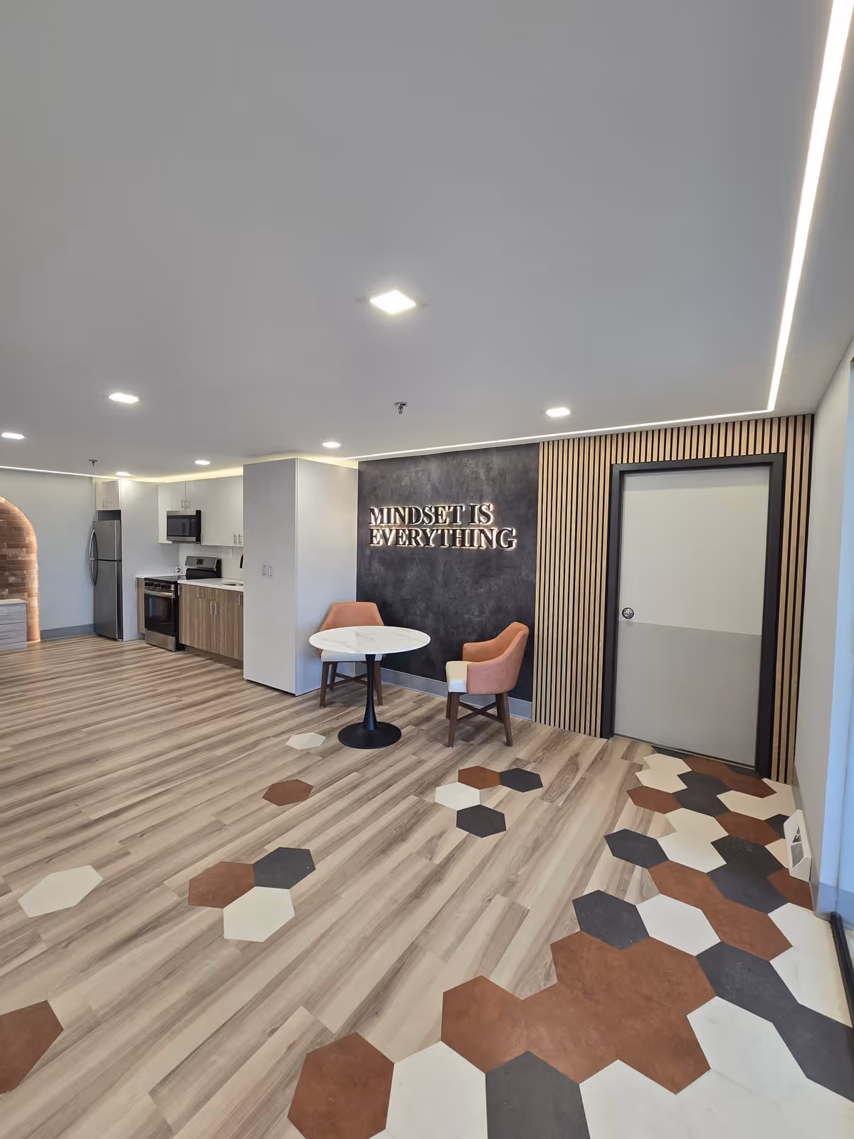 Modern interior space with a small round table and two pink chairs against a wall with the phrase 'MINDSET IS EVERYTHING' displayed. The room features a kitchenette with a refrigerator, microwave, and stove. The floor has a unique design with hexagonal tiles in brown, black, and cream colors blending into wood-patterned flooring. The ceiling has recessed lighting and a strip of LED lights along the edge.