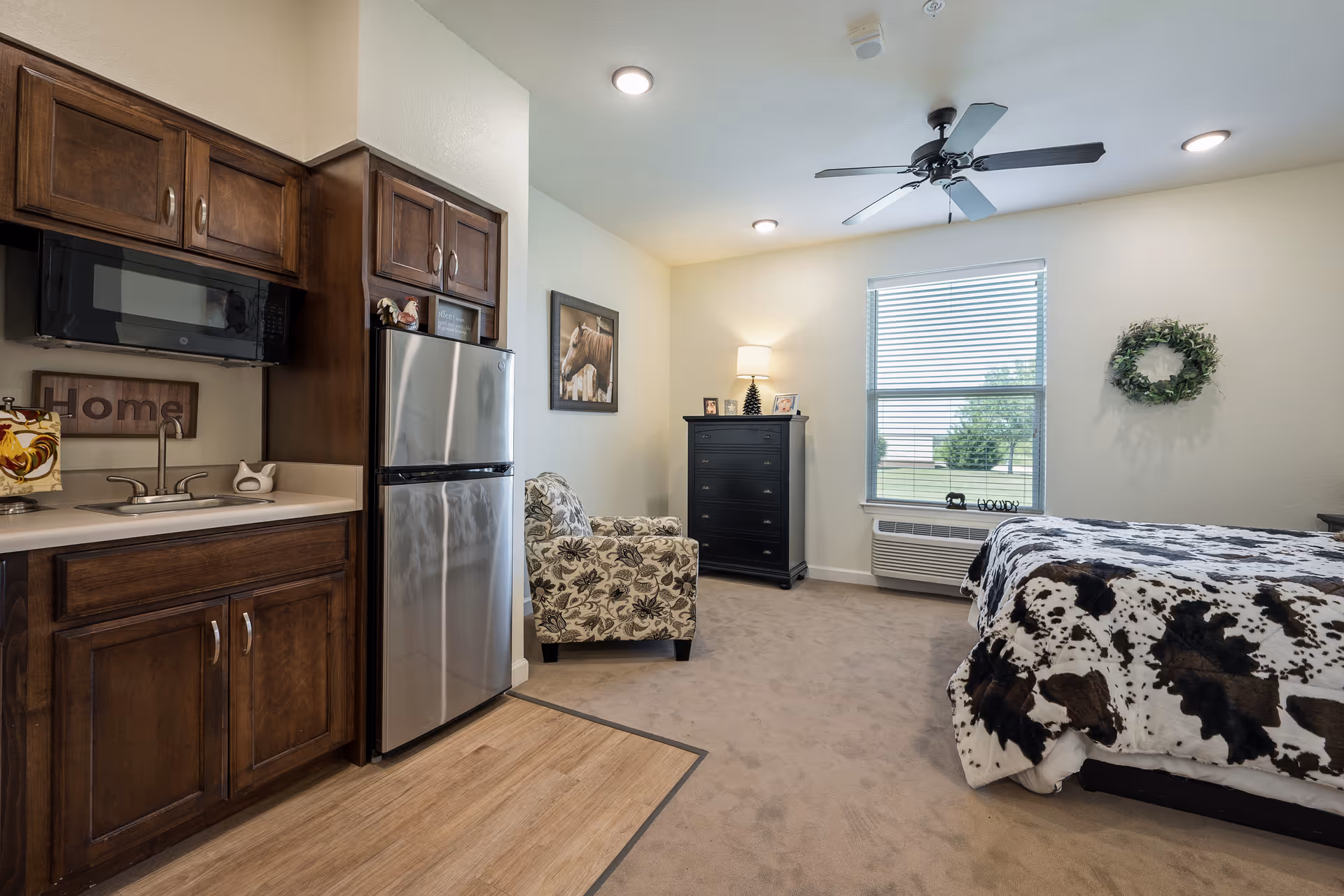 Studio-style resident room with a kitchenette featuring wooden cabinets and a stainless refrigerator, an armchair, dresser, window, and a bed with a cow-print blanket.