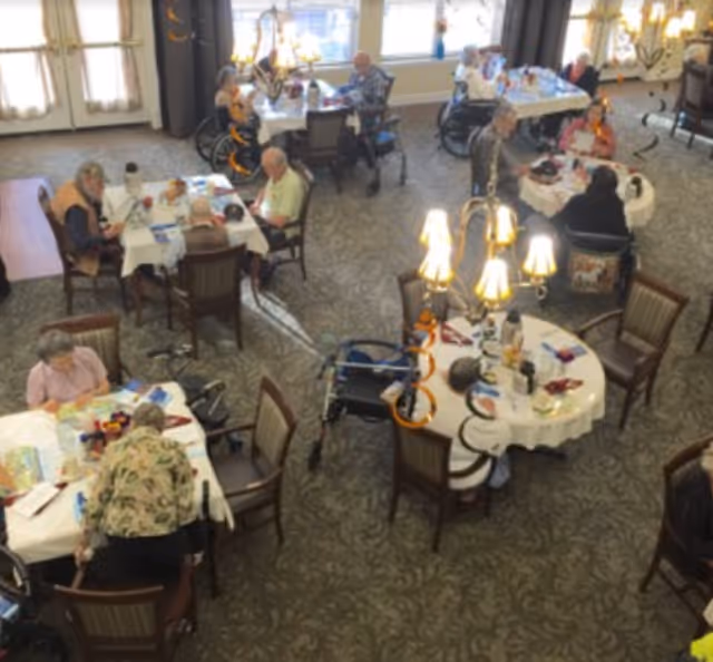 Overhead view of seniors seated at round dining tables in a carpeted assisted-living dining room.