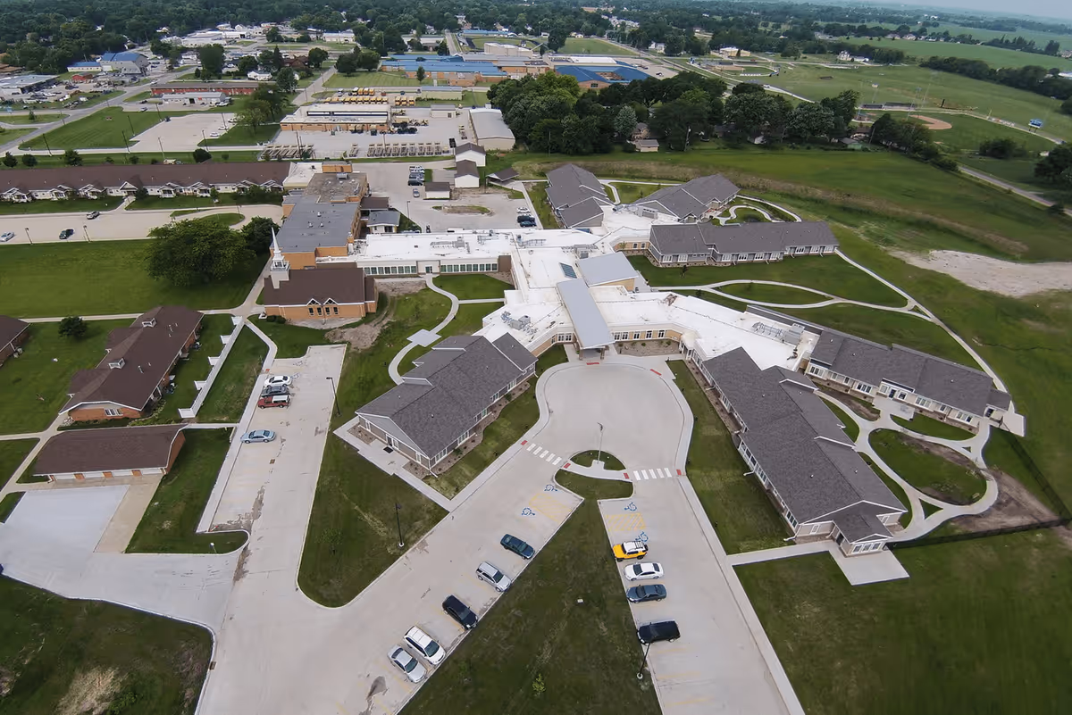 Aerial view of Eden Acres Campus of Perry Lutheran Homes showing multiple connected buildings with gray roofs, surrounding green lawns, parking lots with several cars, and paved walkways. The campus is situated in a semi-rural area with fields and trees in the background.