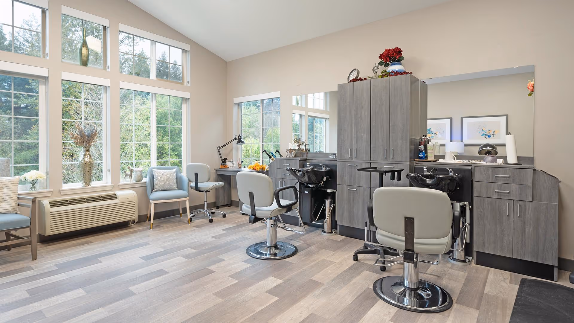 A bright and spacious salon area with large windows letting in natural light. The room features two salon chairs in front of a large mirror and cabinetry, along with additional seating and a desk by the windows. The floor is light wood, and the walls are painted a soft beige.