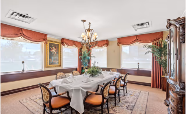 A formal dining room with a rectangular table covered with a white tablecloth, set with folded napkins and glassware. The room has eight upholstered chairs with wooden frames, large windows with red valances and curtains, a chandelier hanging above the table, a framed floral painting on the wall, and a wooden china cabinet on the right side.
