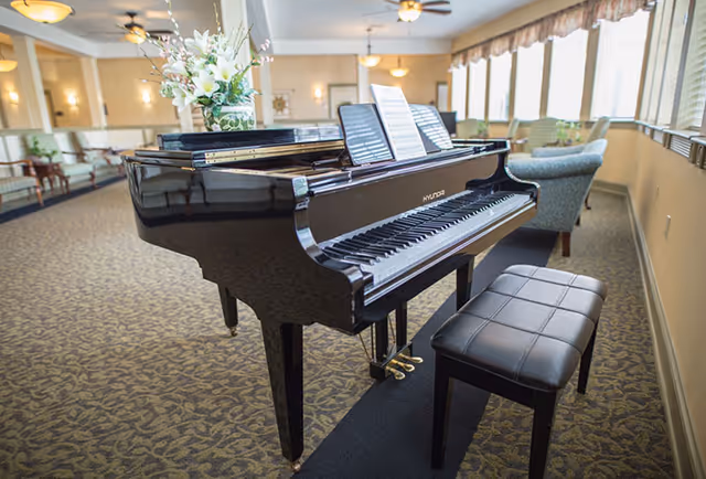 A black grand piano with sheet music on the stand and a flower arrangement on top, placed in a spacious room with patterned carpet, beige walls, ceiling fans, and large windows letting in natural light. There are upholstered chairs and sofas arranged along the walls.