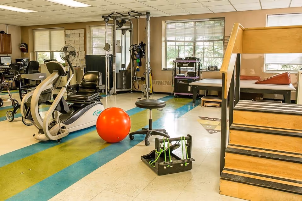 A bright rehabilitation gym with a recumbent bike, red therapy ball, pulley machine, exercise stairs and therapy tables.