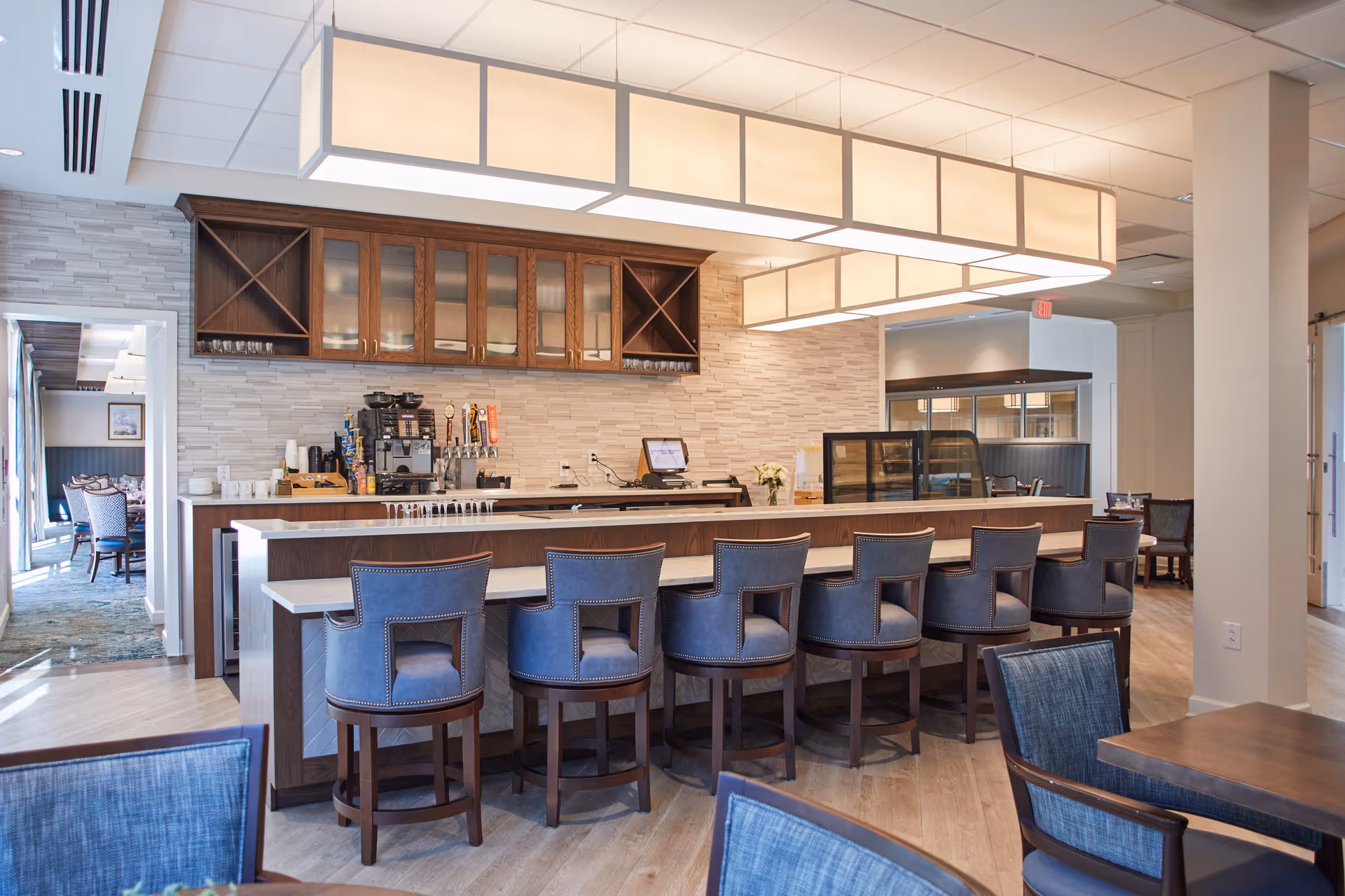 A modern dining area with a long counter and seven blue upholstered bar stools. Behind the counter are wooden cabinets with glass doors, a coffee machine, and several beverage taps. The room features light wood flooring, a large rectangular ceiling light fixture, and additional seating with blue chairs and wooden tables in the foreground.