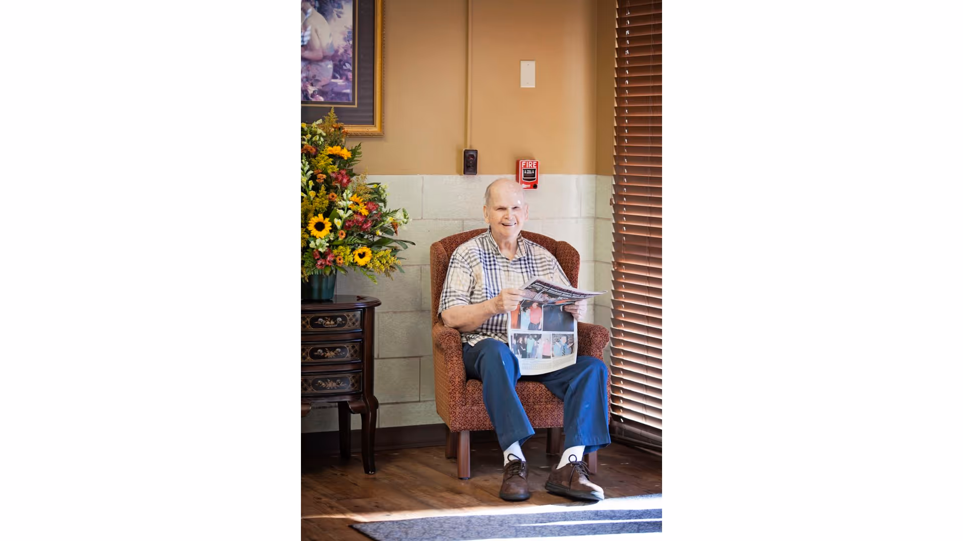 An elderly man sitting in a patterned armchair near a window with wooden blinds, holding and reading a newspaper. Next to him is a small wooden table with a large colorful flower arrangement. The room has beige walls with a fire alarm and a framed picture partially visible.