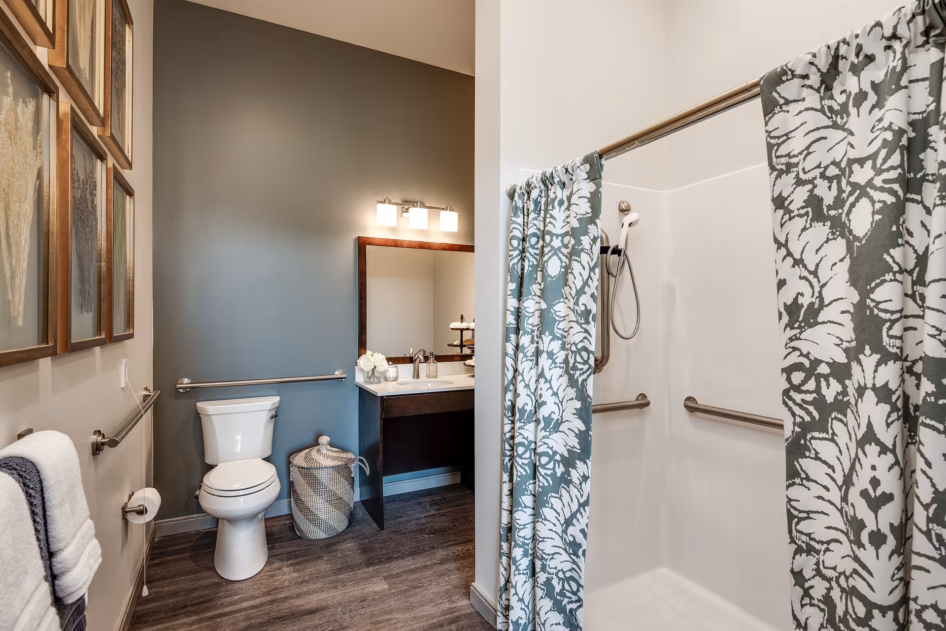 A modern bathroom featuring a white toilet, a vanity with a sink and mirror, a shower with a floral-patterned curtain, and several framed artworks on the wall. The bathroom has grab bars near the toilet and inside the shower for accessibility.