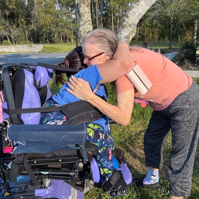 An elderly woman in a wheelchair with purple cushions is being hugged by another woman wearing a coral shirt and gray pants outdoors near a tree and grassy area.