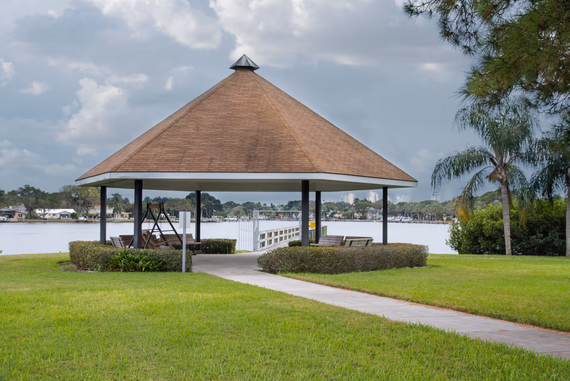 A wooden gazebo with a brown shingled roof situated on a grassy area near a body of water. There are benches under the gazebo and a paved walkway leading to it. Trees and houses are visible in the background across the water under a cloudy sky.