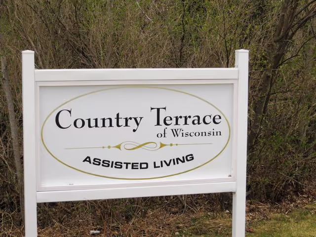 A white outdoor sign with gold and black lettering that reads 'Country Terrace of Wisconsin ASSISTED LIVING' set against a background of leafless bushes and grass.