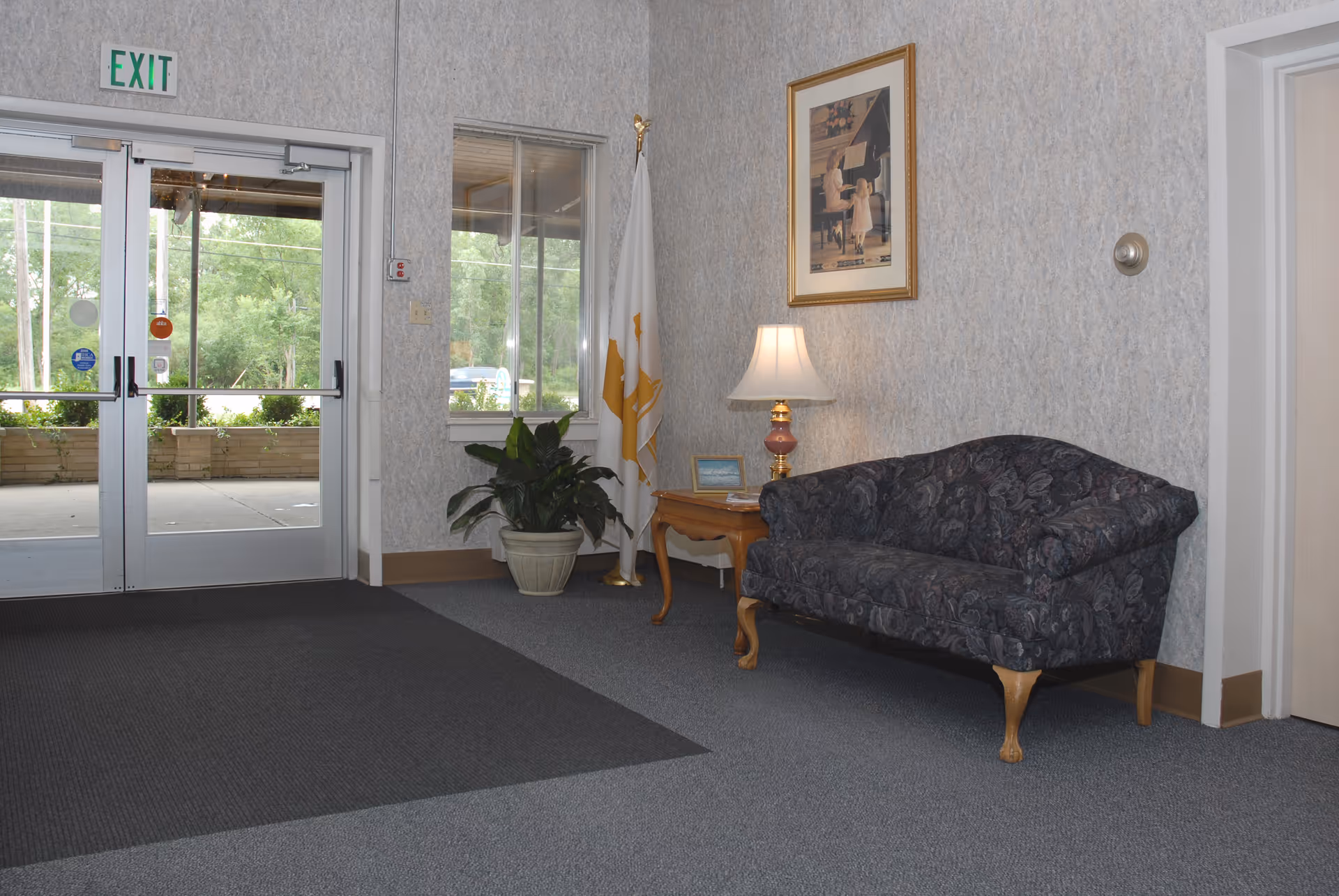 Interior view of a facility entrance area with double glass doors marked with an exit sign. There is a dark patterned couch, a wooden side table with a lamp and a small framed picture, a potted plant, and a flag in the corner. A framed picture hangs on the wall above the couch.