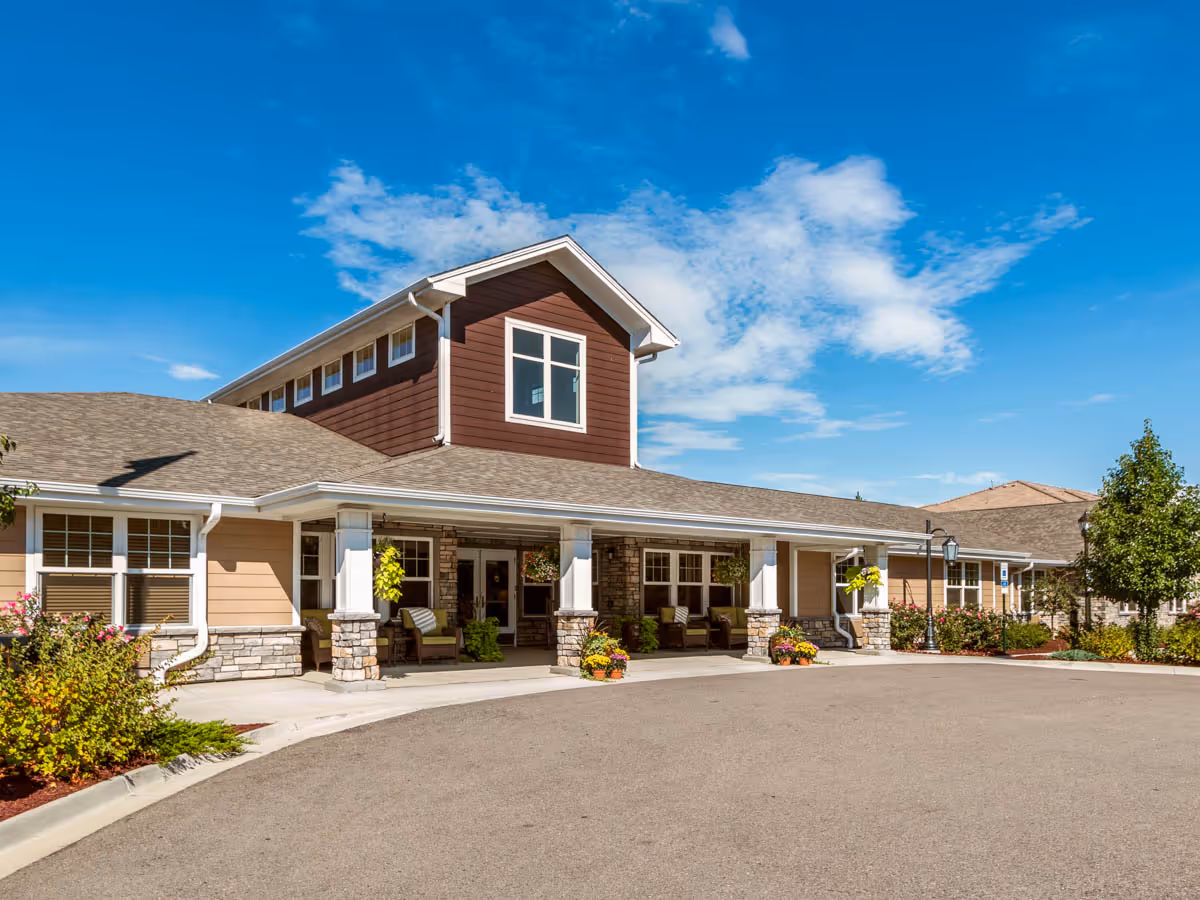Front exterior of a single-story senior living building with a covered entrance supported by stone columns, landscaped flower beds, and a paved driveway.