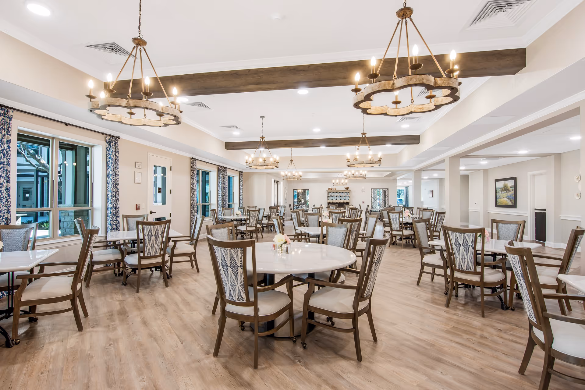 Bright senior dining room with round tables, wooden chairs, chandeliers, and large windows.
