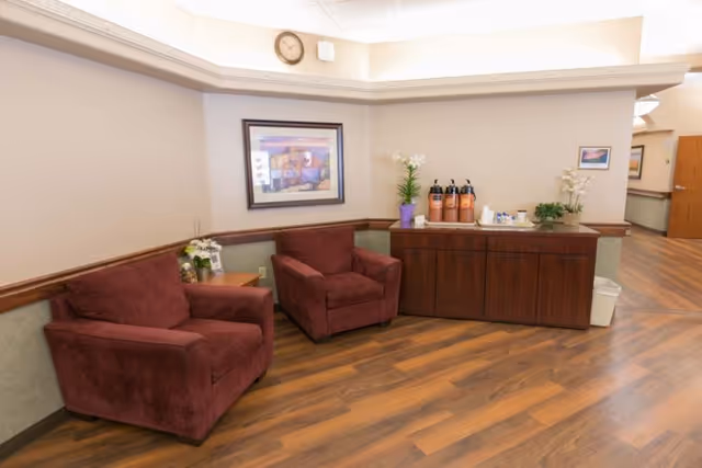 A small lounge area with two maroon armchairs, a side table, framed artwork, and a wooden cabinet holding coffee dispensers against a hallway wall.