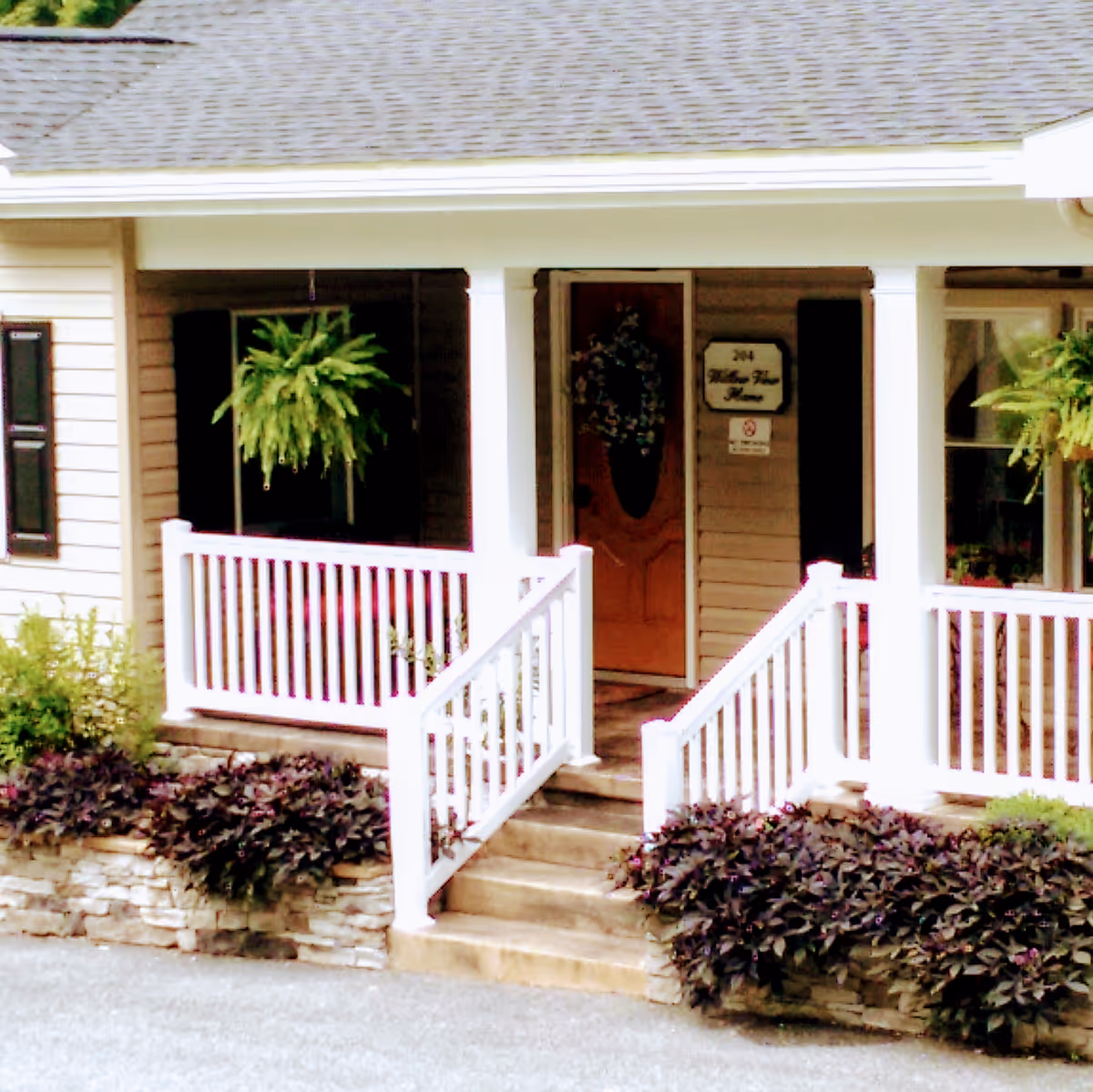 Front porch entrance of a building with white railings, steps leading up to a wooden door decorated with a wreath. There are hanging green plants on either side of the porch and a sign next to the door that reads '204 Willow View Home'.