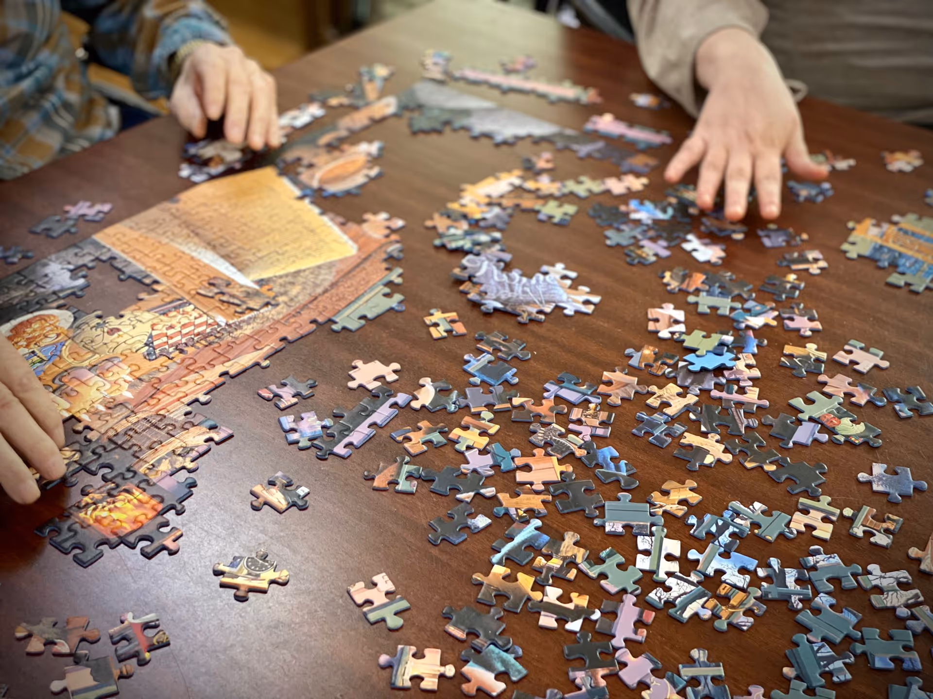 Two people working together on assembling a jigsaw puzzle on a wooden table, with many puzzle pieces scattered around.