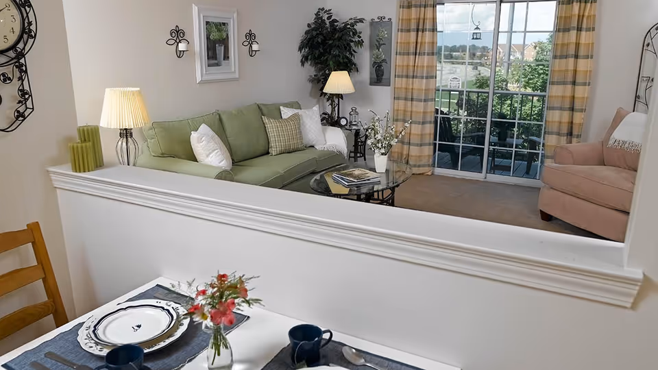 Bright living room with a green sofa, glass coffee table, armchair, sliding glass doors to a balcony, and a small dining place setting in the foreground.