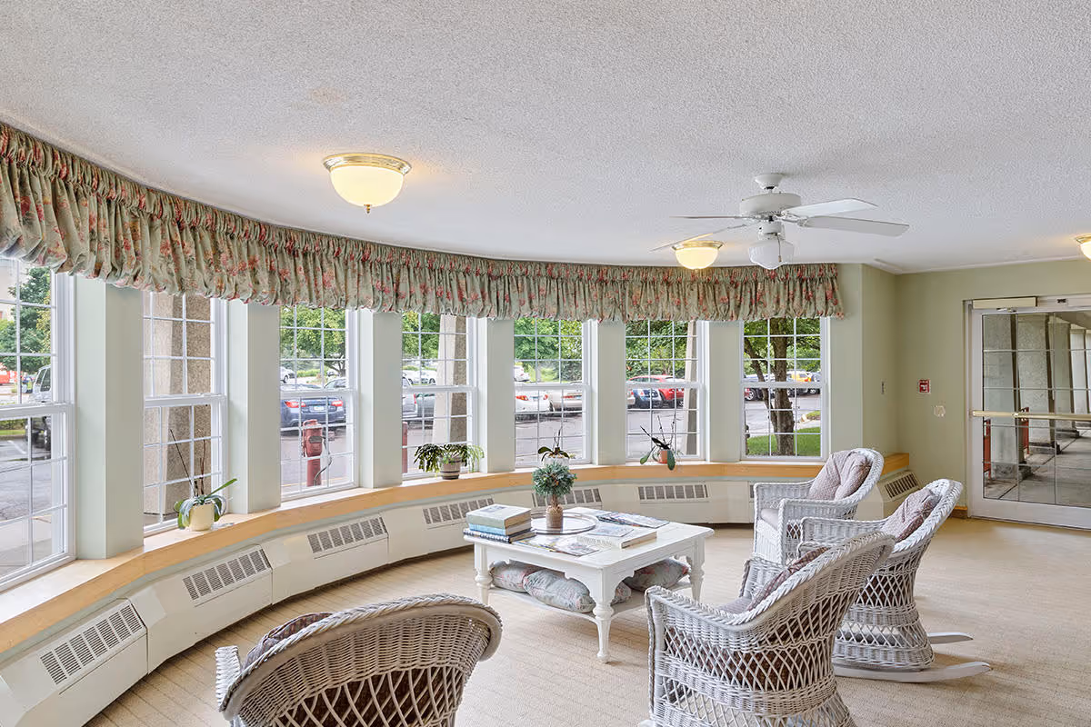 A bright and cozy senior living facility common area with large curved windows letting in natural light. The room features wicker rocking chairs with cushions arranged around a white coffee table that holds books and a small plant. Floral valances adorn the windows, and there are potted plants on the window ledge. The space has beige carpeting, ceiling lights, and a ceiling fan. A glass door leads to another area of the building.