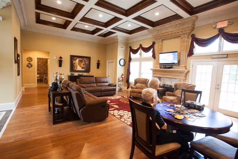 A spacious living room with wooden flooring, a coffered ceiling, and beige walls. The room features a large brown sectional sofa, two armchairs near a fireplace with an ornate mantel, and a wall-mounted TV above the fireplace. Two elderly individuals are seated at a wooden table working on a puzzle. Large windows with burgundy curtains and glass doors allow natural light into the room.