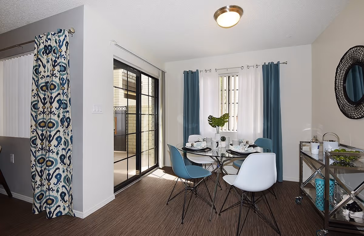 A bright dining area with a round glass table set for four, featuring blue and white chairs. The room has large windows with blue and white curtains, a sliding glass door leading outside, and a metal serving cart with decorative items and a round mirror on the wall.