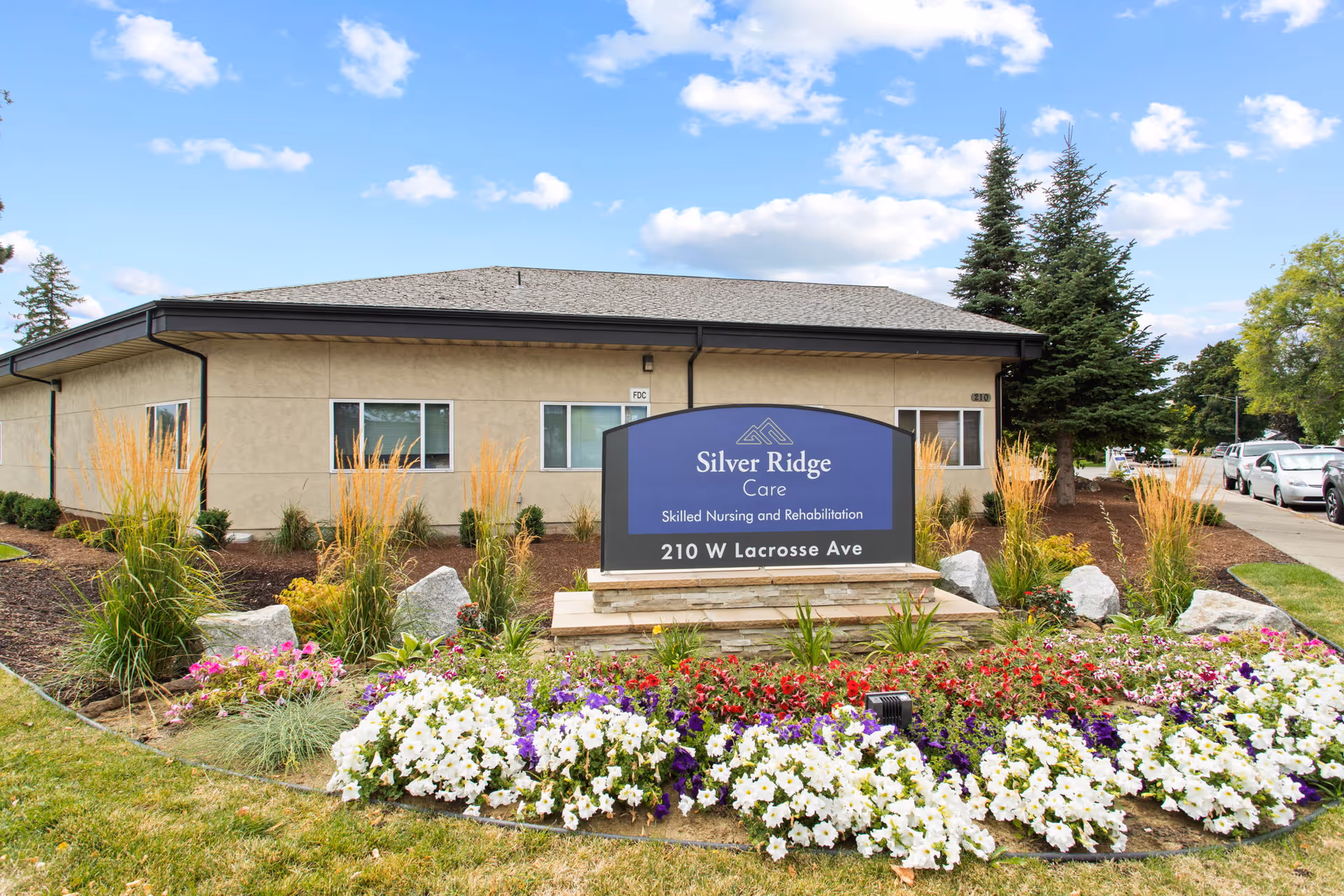 Exterior view of Silver Ridge Care facility showing a single-story building with beige walls and a gray roof. In front of the building is a landscaped garden with colorful flowers and ornamental grasses. A large blue sign in the garden reads 'Silver Ridge Care Skilled Nursing and Rehabilitation 210 W Lacrosse Ave'. Several cars are parked along the street to the right.