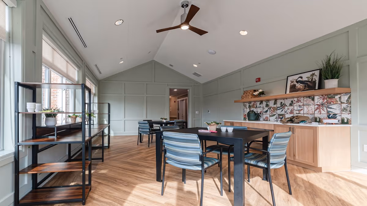 A bright and spacious room with light green paneled walls and wood flooring. The room features a black table with four striped cushioned chairs, a ceiling fan with wooden blades, and large windows letting in natural light. There are open shelves on the left side with small potted plants and cups, and a wooden cabinet with a decorative backsplash and plants on the right side.