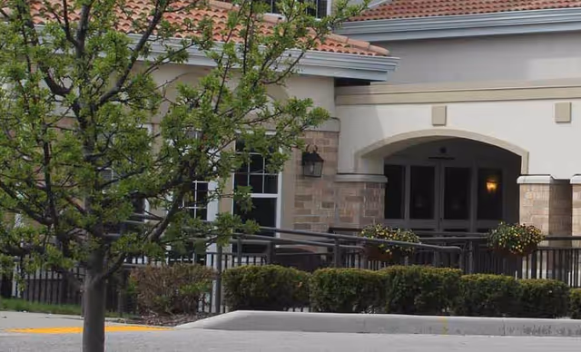 Exterior view of a building with beige and brick walls, a tiled roof, and an arched entrance. There is a tree with green leaves and trimmed bushes in front of the building.