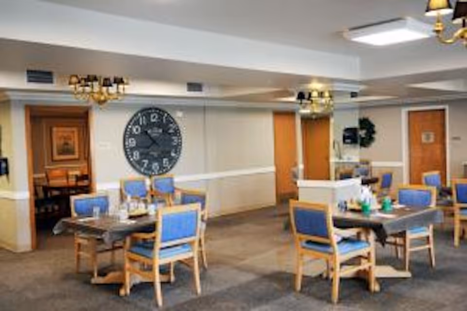 Dining room with wooden tables and blue-upholstered chairs, a large wall clock, and overhead chandeliers.