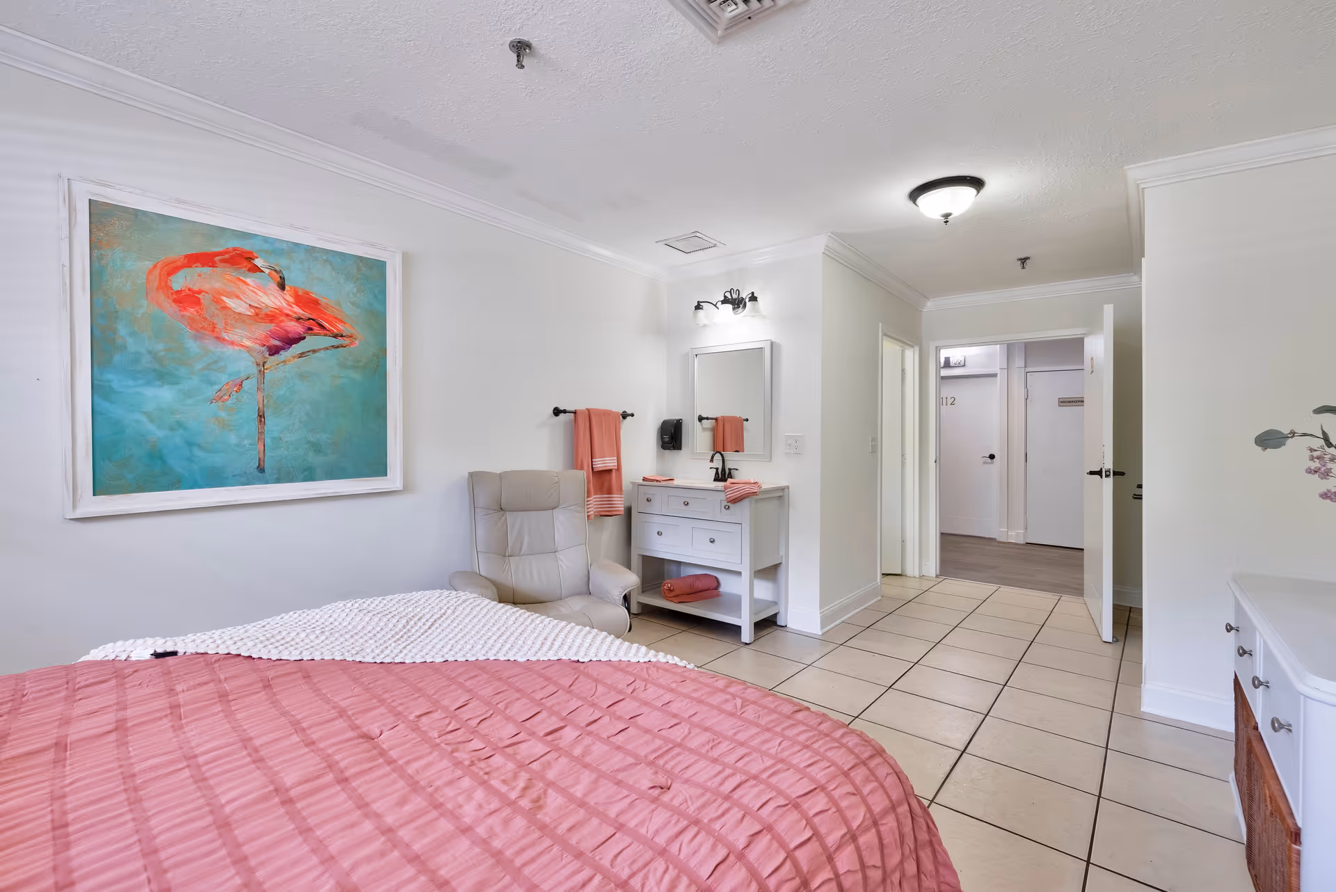 A bright bedroom with a pink bedspread, a beige recliner chair, a white vanity with a mirror and pink towels, and a large framed painting of a flamingo on the wall. The room has tiled flooring and white walls, with an open door leading to a hallway.