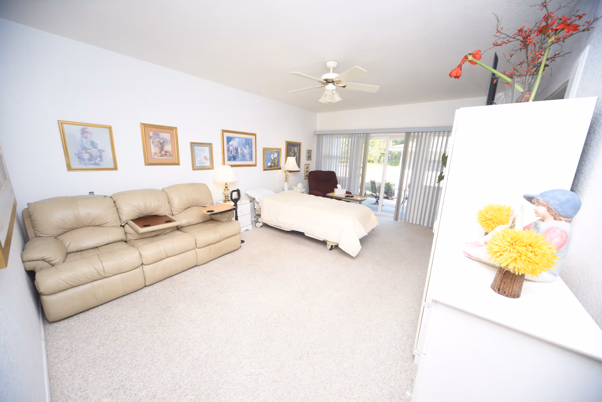 A bright room featuring a beige leather sofa with two wooden trays, a hospital-style bed with white bedding, a maroon recliner chair, and a white dresser with decorative items including a figurine and yellow flowers. The walls are adorned with framed artwork, and large sliding glass doors with vertical blinds open to an outdoor patio area.