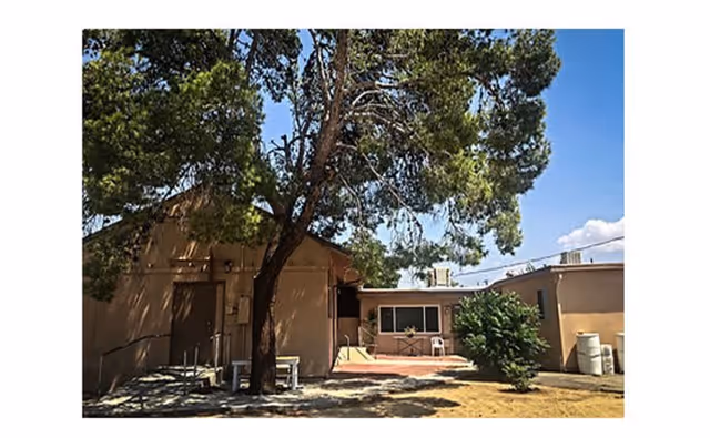 Exterior view of a single-story building with beige walls, surrounded by trees and a small yard with dry grass under a clear blue sky.