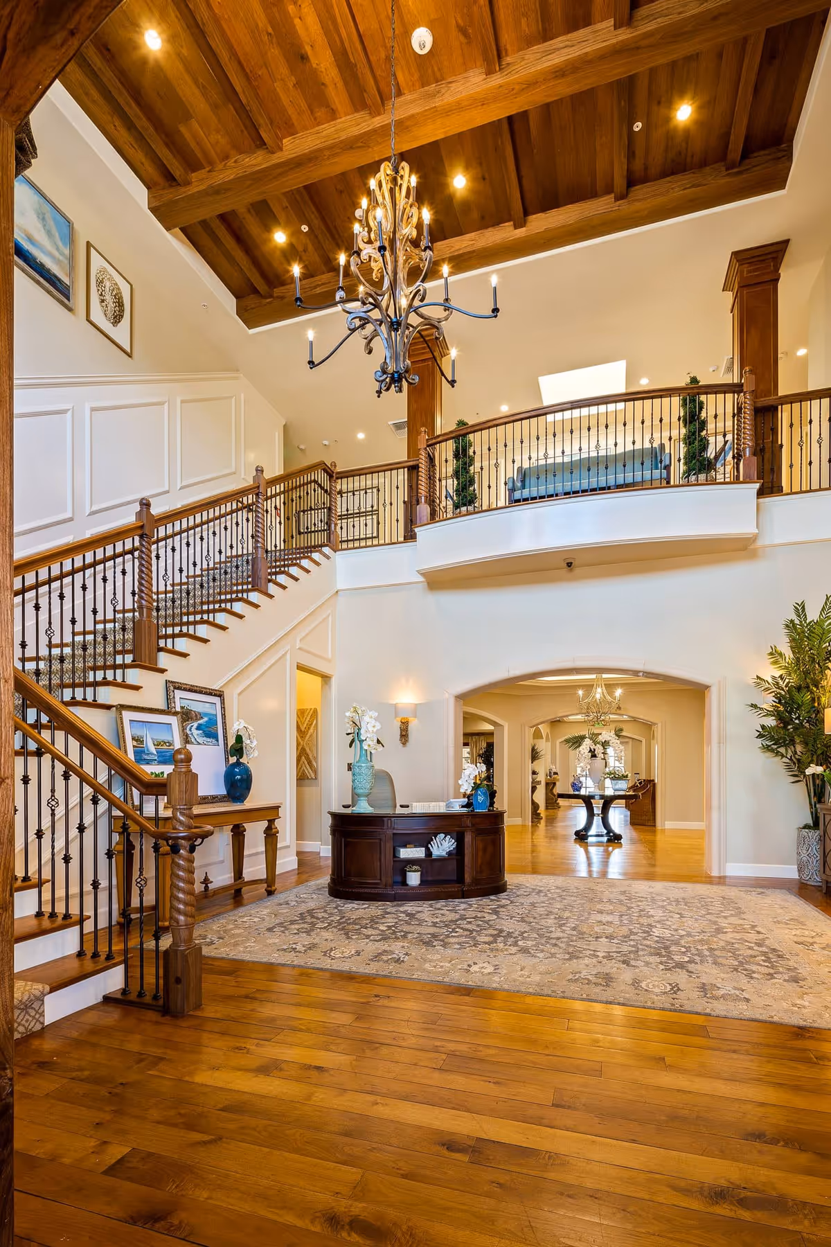 Spacious and elegant interior of The Kensington Redondo Beach featuring a wooden staircase with wrought iron railings, a large chandelier hanging from a wooden ceiling, a round wooden reception desk with decorative vases and flowers, framed artwork on the wall, and a patterned area rug on polished hardwood floors. The background shows an archway leading to another room with more decorative elements and furniture.