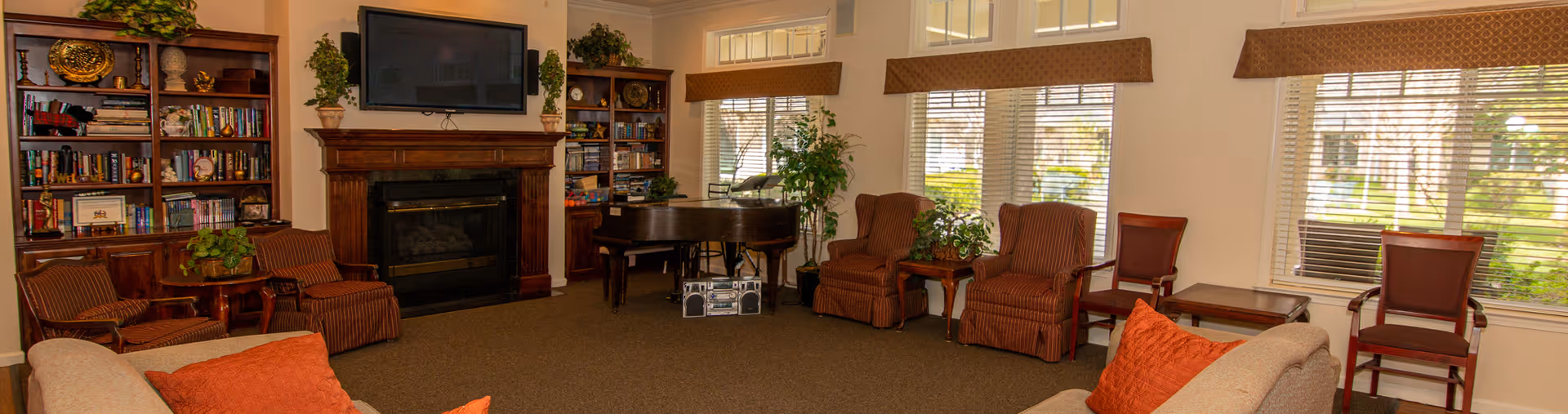 Communal living room with sofas and armchairs arranged around a fireplace and TV, bookshelves, piano, and large windows.