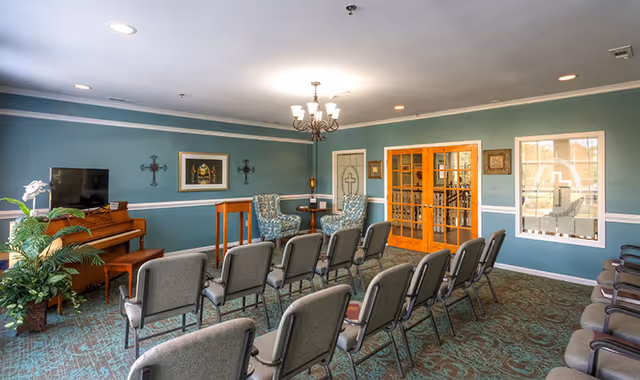A well-lit room with rows of gray cushioned chairs arranged facing a piano and two upholstered armchairs with a small round table between them. The walls are painted teal with white trim, decorated with framed artwork and crosses. There are double wooden doors with glass panes and a window with a cross design on the right side. A chandelier hangs from the ceiling.