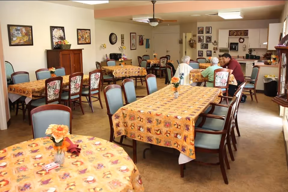 A senior living facility dining room with several tables covered in patterned tablecloths and decorated with small flower vases. Three elderly individuals and a caregiver are seated at one table near the kitchen area. The room is warmly lit with wall art and a clock on the walls.