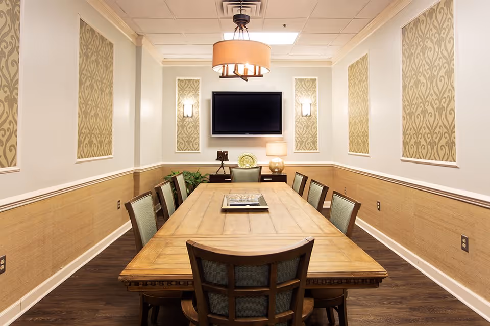 A well-lit conference or dining room with a long wooden table surrounded by eight chairs. The walls are decorated with patterned fabric panels and two wall sconces. A flat-screen TV is mounted on the far wall above a sideboard with a lamp and decorative items. A ceiling light fixture hangs above the table.